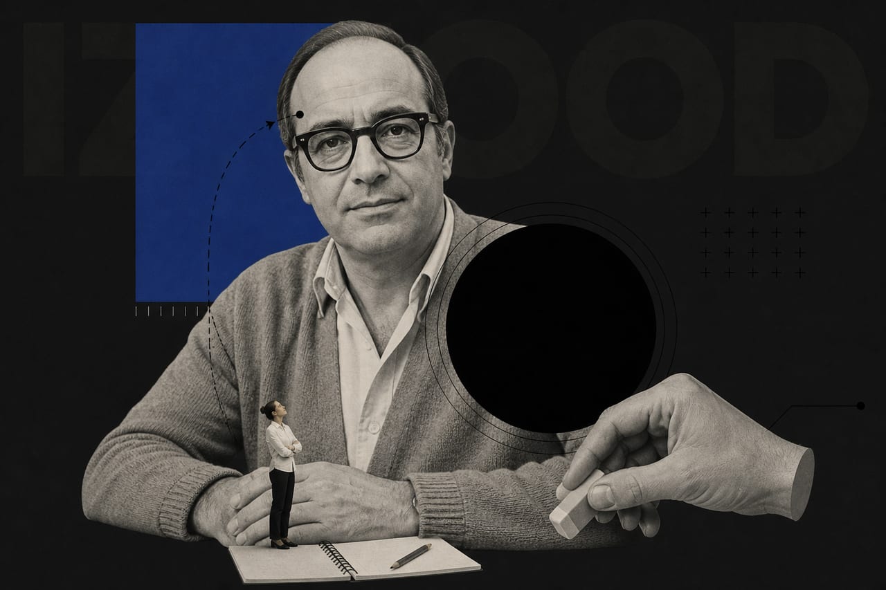 A large black-and-white photograph of a middle-aged man in a rumpled cardigan and thick-framed glasses fills most of the frame, seated at a plain wooden desk.